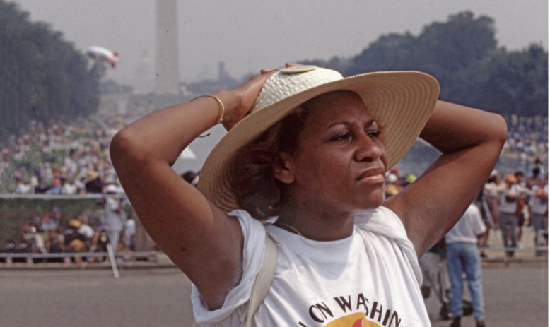Photo of Black woman wearing straw hat and t-shirt reading "March on Washington," with her hands on her head looking away from the camera, standing in front of the Washington Monument