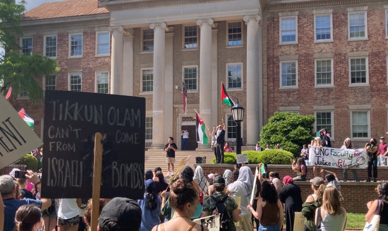 From-behind image of protestors in front of UNC's Wilson library. Two signs are visible: "Tikkun Olam can't come from Israel's bombs" and "UNC Faculty for Justice in Palestine." Speakers at the front hold Palestinian flags.