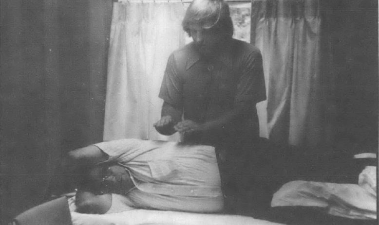 Black and white photo of man, standing, holding his hands over another man lying on his side on a table