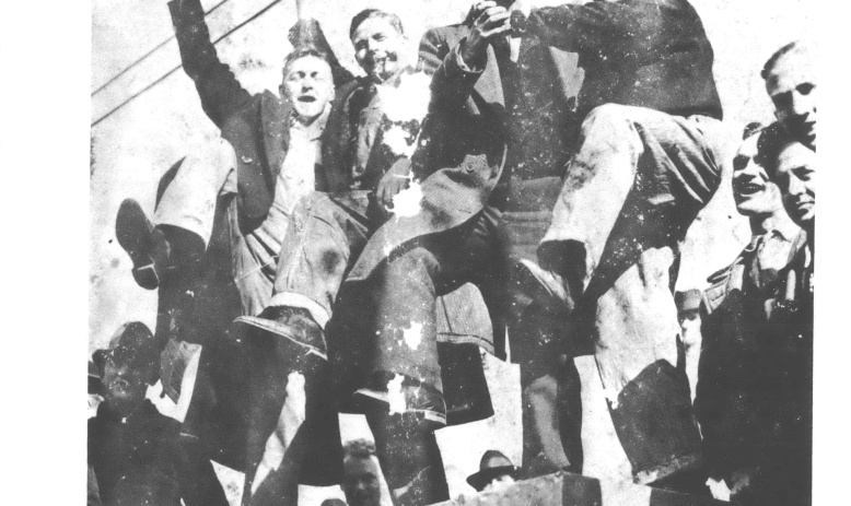 Black and white photo looking up at a group of man standing on a wall, some with fists and hats raised