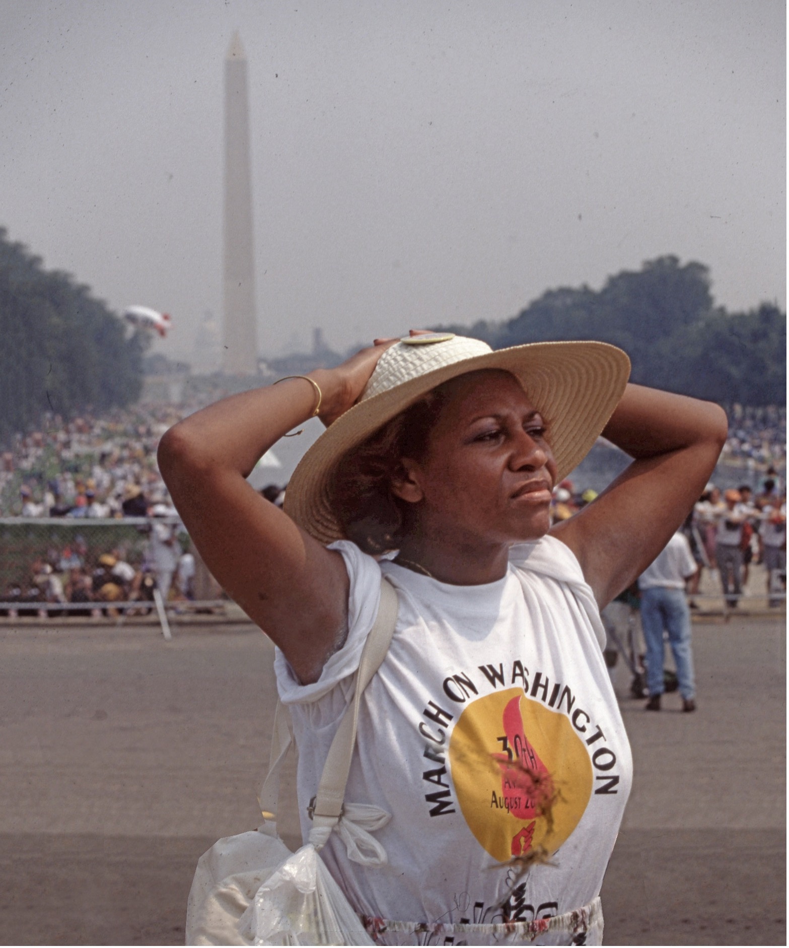 Photo of Black woman wearing straw hat and t-shirt reading "March on Washington," with her hands on her head looking away from the camera, standing in front of the Washington Monument