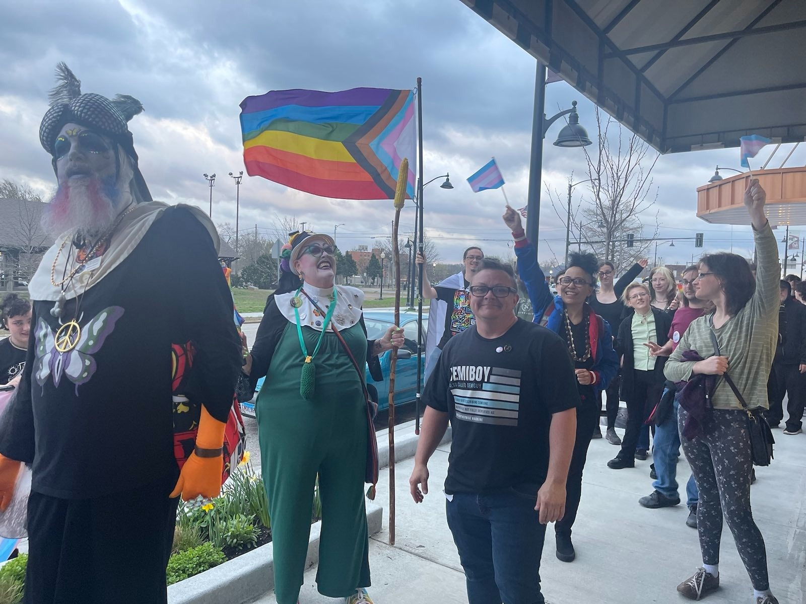 The image depicts a group of individuals smiling, holding gay and trans pride flags, and walking along a sidewalk in a parade on Trans Day of Visibility.