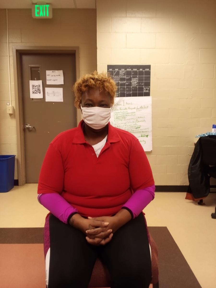 Black woman in a face mask, red shirt, and black pants, sitting in a chair and looking at the camera