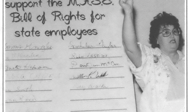 woman stands with her fist raised in front of a large board that reads "We the undersigned support the M.A.S.E. Bill of Rights for state employees" 