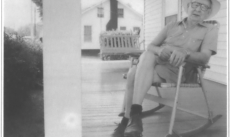 retired forman sits on a porch in a rocking chair wearing a hat 