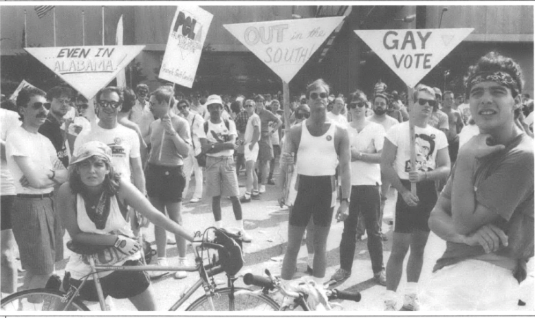 Lots of people in front of a building, in the middle ground people holding 4 signs that read "...Even in Alabama!" 'OUT in the SOUTH" "Gay Vote" in the fore ground two bikers one femme and one masc apparently waiting around looking out past the camera
