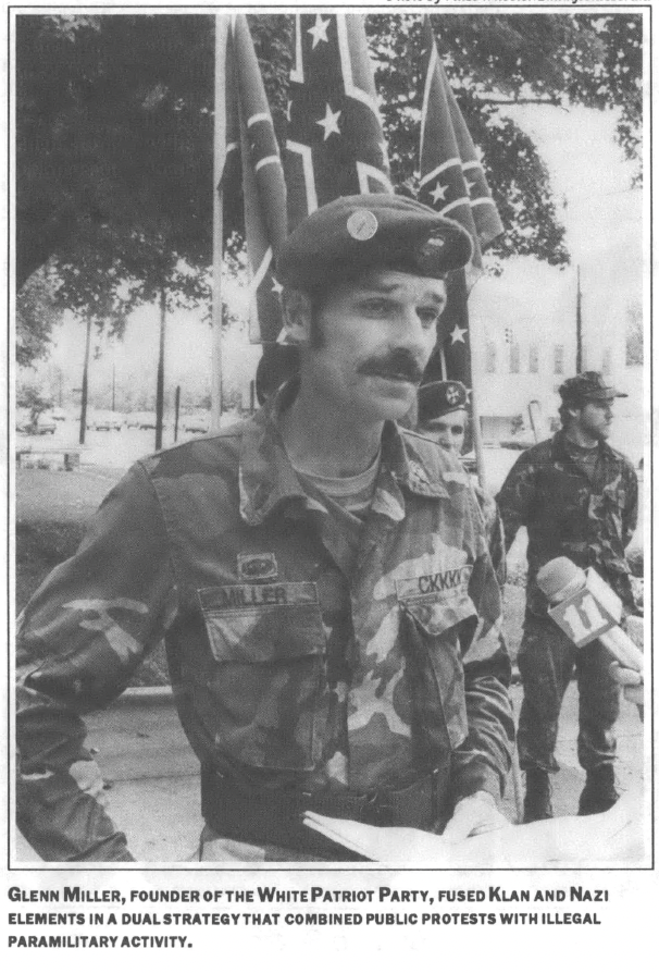 Photo of Glen Miller, founder of the White Patriot Party, talking to a television news reporter, wearing military clothes, a baret, and standing between two other men, three confederate flags, and a tree, at a park 