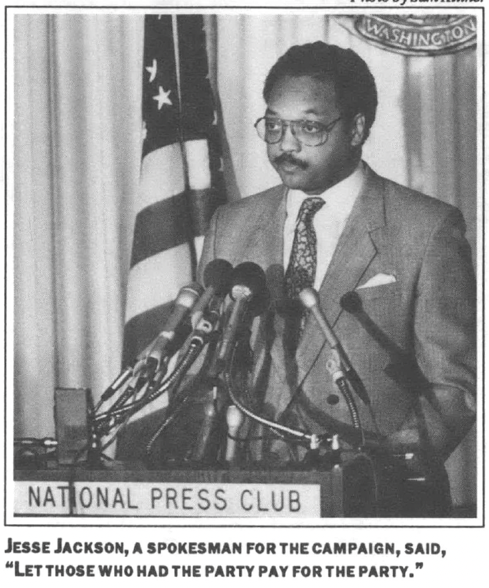 Man in a suit with glasses standing at a podium with 6 mics giving a press conference in front of a national seal and american flag