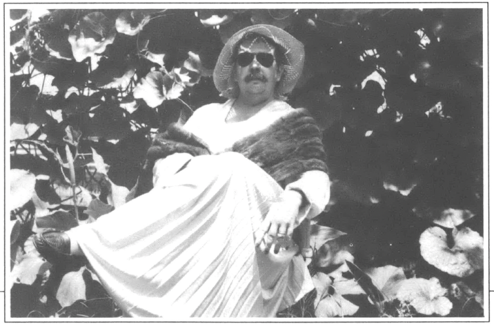 Black and white photograph of a masc person with a mustache, straw hat, and sunglassess wearing a mink coat over a dress in front of a bush 