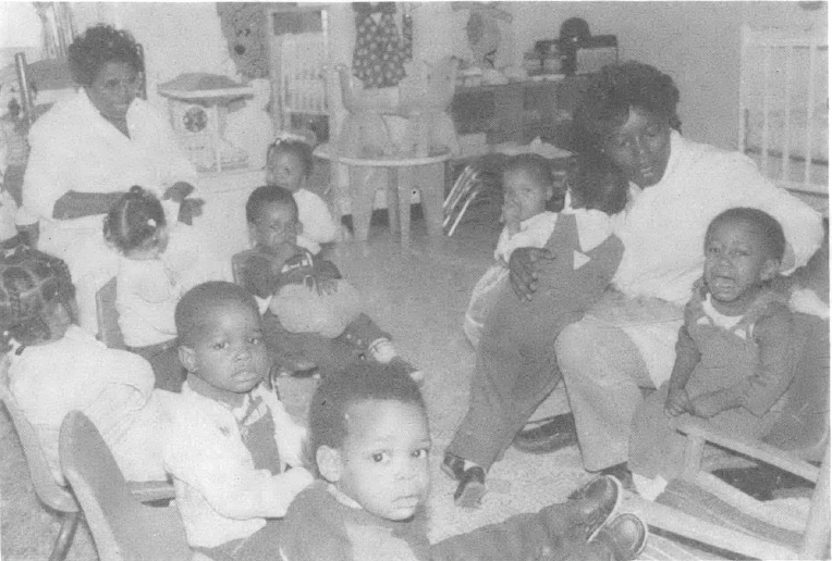 Two teachers and several children sit on the floor of a daycare center