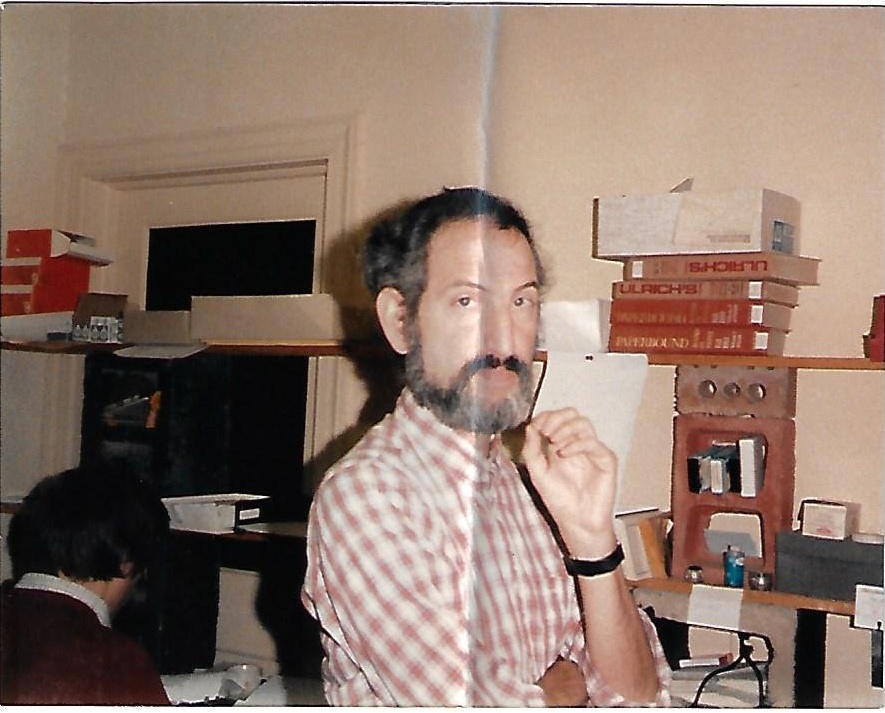 Scanned photo with crease through the middle showing man standing in an office with his hand to his chin, wearing a red and white checked shirt