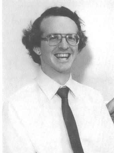 Photo of white man with glasses in collared shirt and tie grinning at the camera 