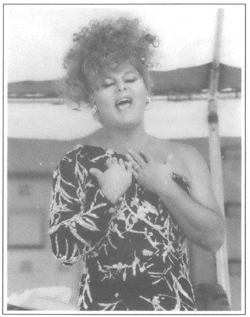 Drag Queen singing at a harbor in single should dress, hands pulled in on her chest, a bay in the background