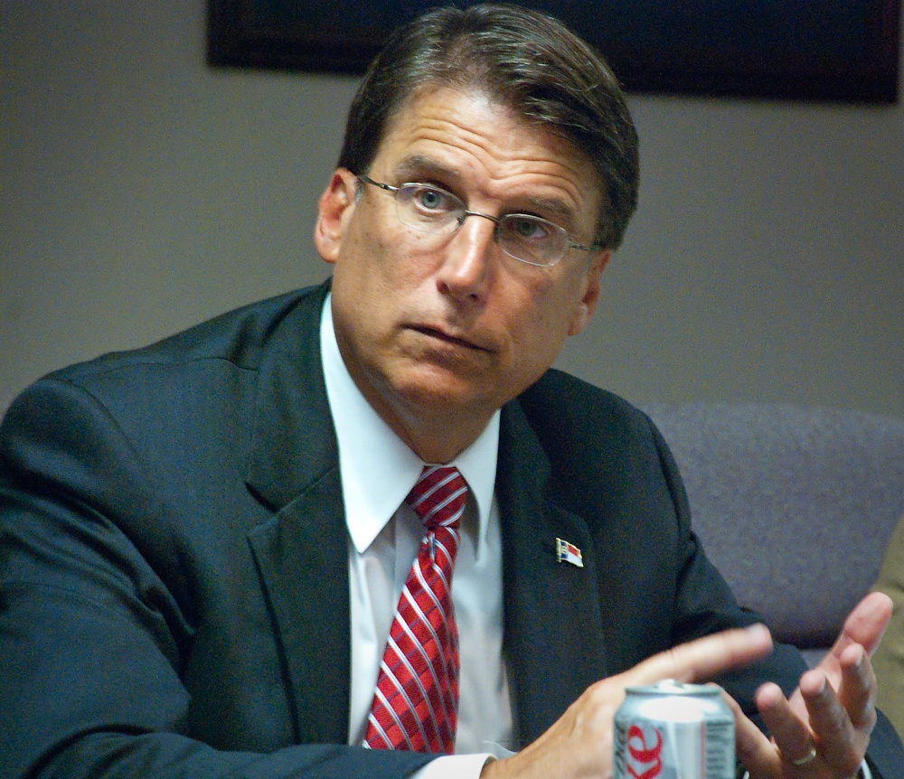 The photo depicts a white man with glasses and brown hair counting on his fingers while looking outside of the camera's view to the upper lefthand corner of the image. He is wearing a dark-colored suit, blue dress shirt, and red tie.