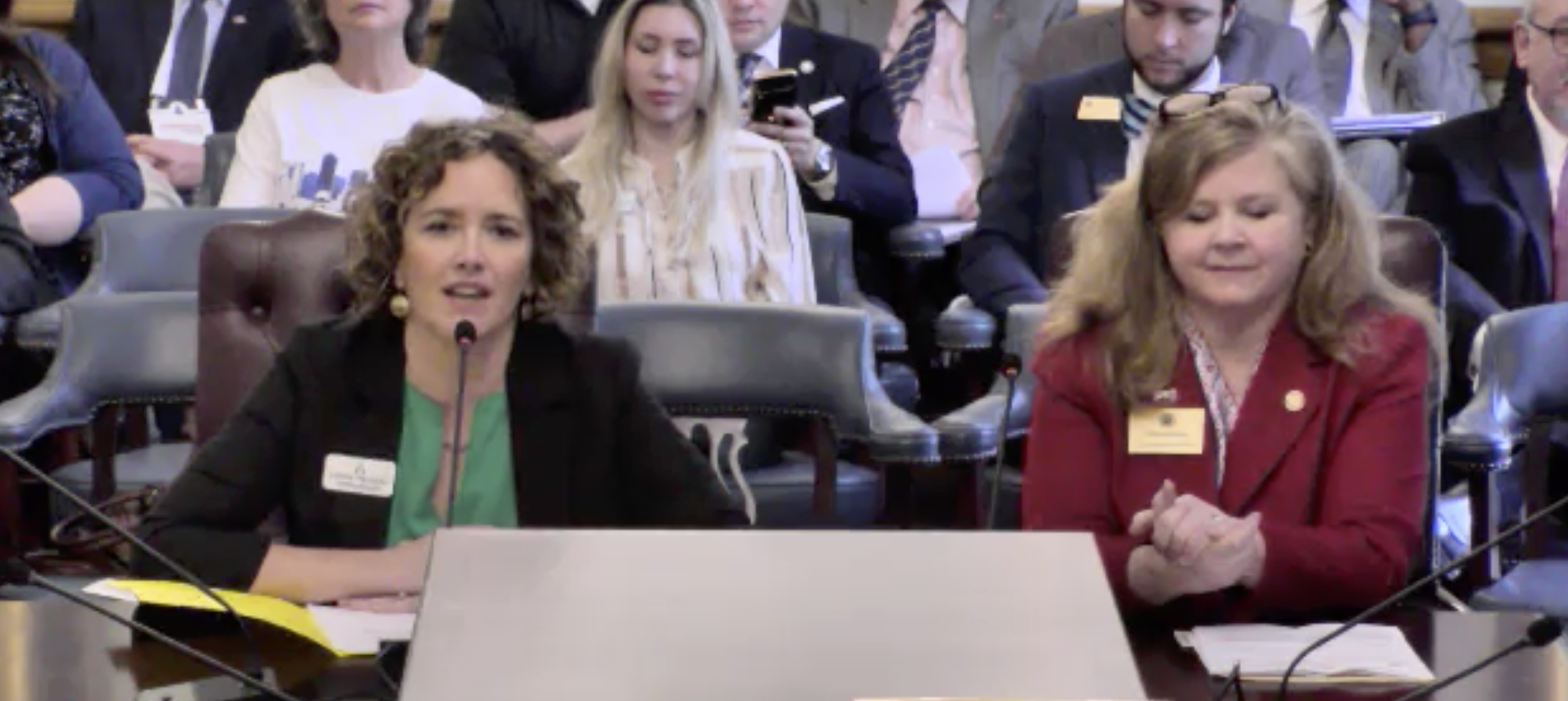 Two women sit in front of microphones at a conference table, with onlookers in the background. The woman on the left is talking.