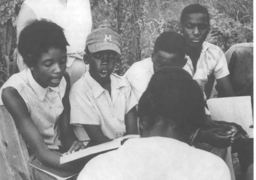 A group of Black students seated outside reading a book. One appears to be reading out loud.