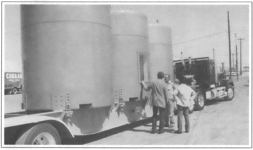 truck rigged to a trailer with three large trailers behind it, three men standing and talking by the middle barrel