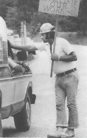 Photo of man in hard hat on picket line holding sign talking to someone in a pickup truck