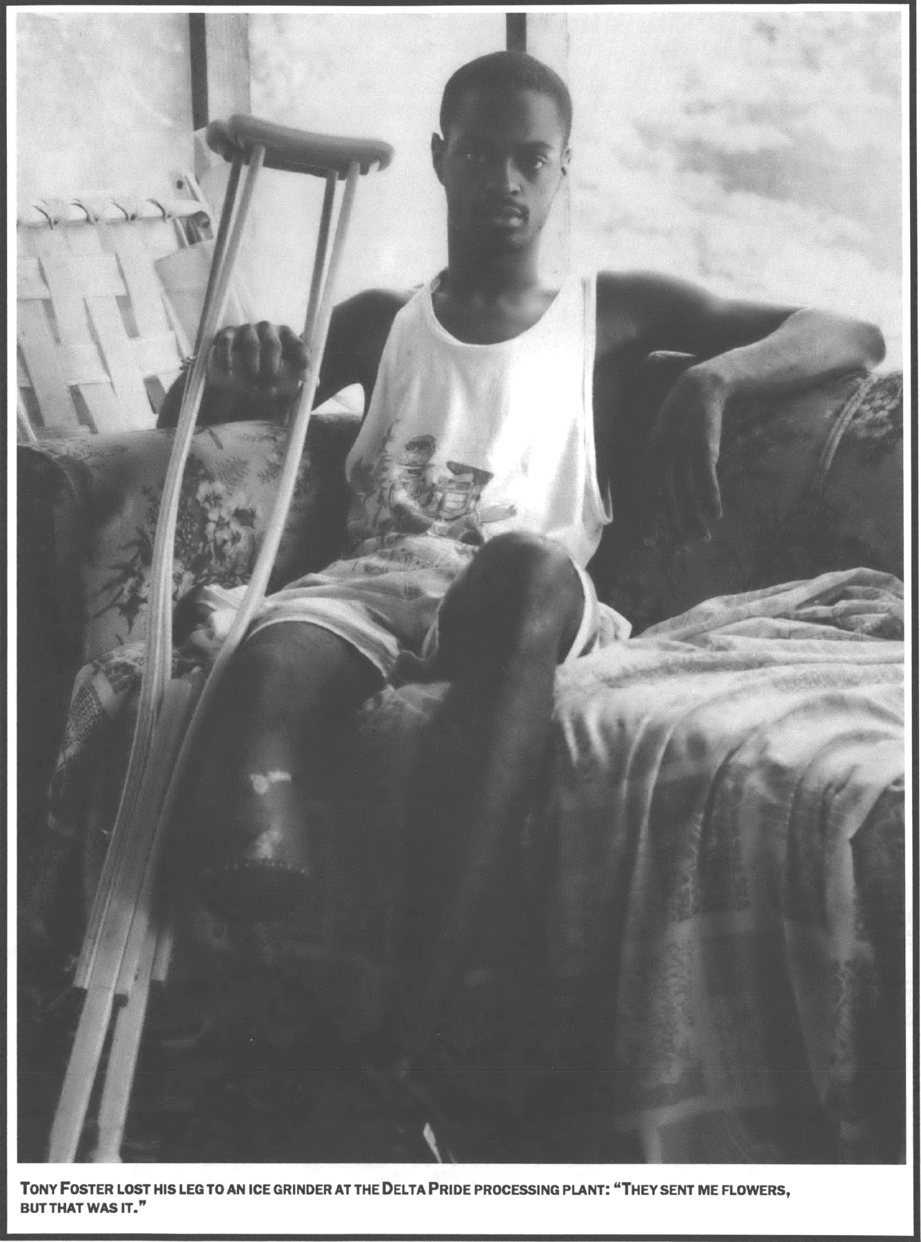 man in tank top seated on a couch by a window facing camera holding a single crutch