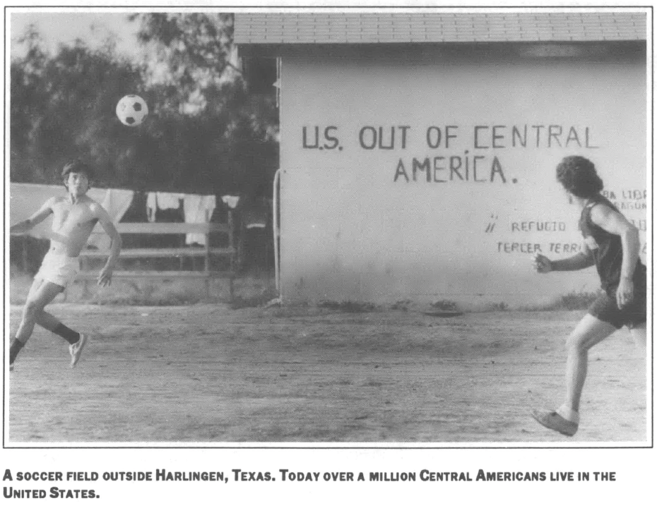 photo of two people playing soccer one shirtless on a dirt field soccerball in the air in front of a clothesline and a building which reads "U.S Out of Central America"