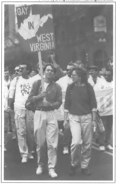 people marching in a street, many wearing white shirts, two people standing in front one holding a sign with the outline of West Virginia on it and "Gay in West Virginia" w