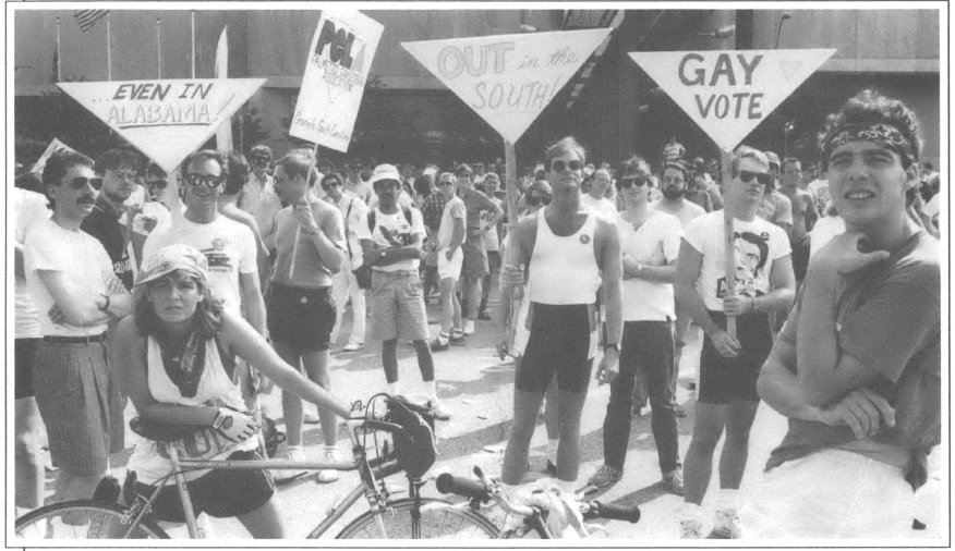 Lots of people in front of a building, in the middle ground people holding 4 signs that read "...Even in Alabama!" 'OUT in the SOUTH" "Gay Vote" in the fore ground two bikers one femme and one masc apparently waiting around looking out past the camera