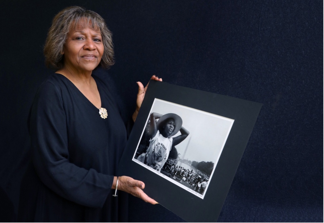 Recent photograph of Raymona Middleton holding a large photograph of herself at the March on Washington thirty years ago, against a black background