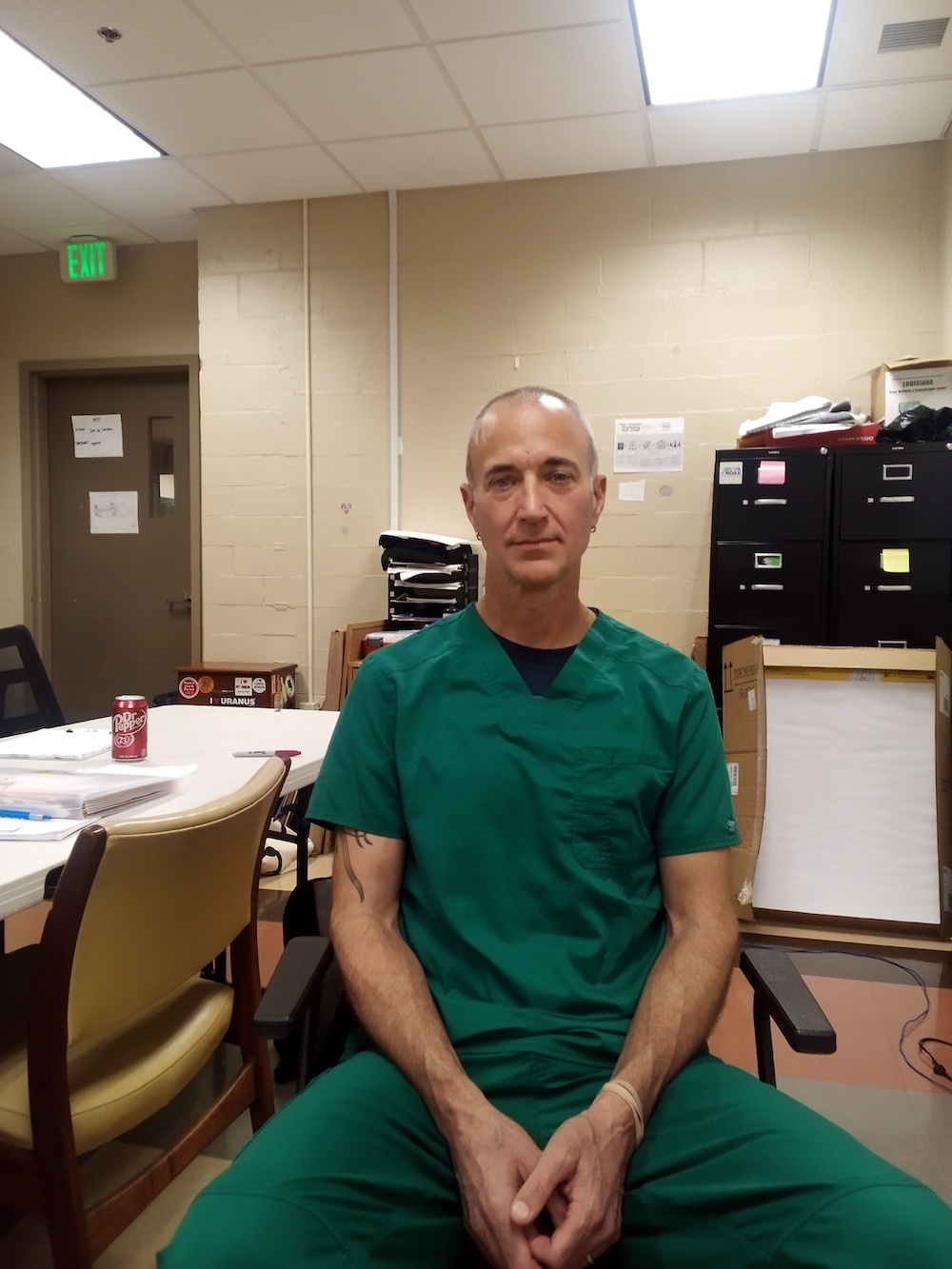 Bald white man in green scrubs, sitting in chair and looking directly at the camera