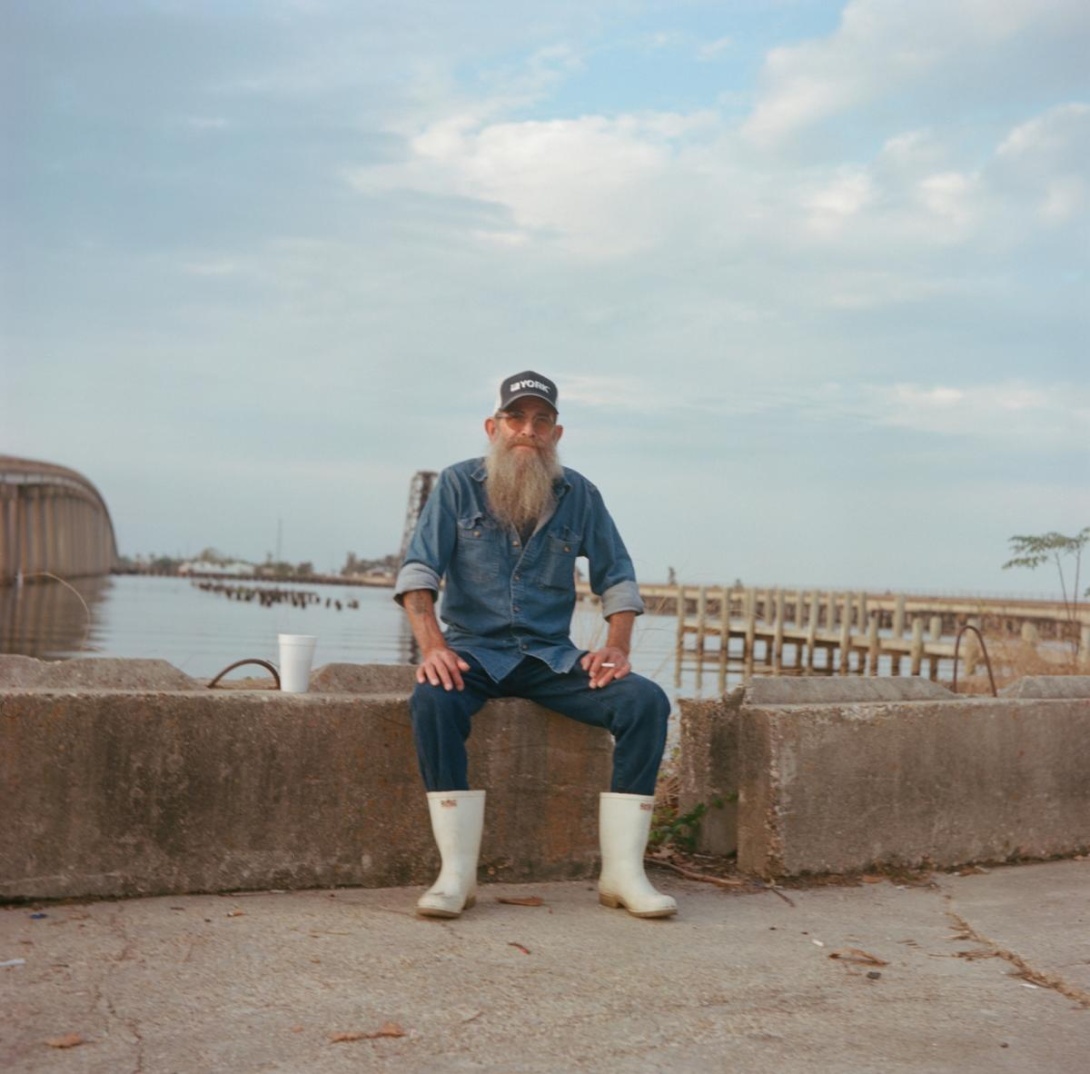 White man wearing ball cap, sunglasses, denim shirt and pants, and white muck boots sitting near lake's edge